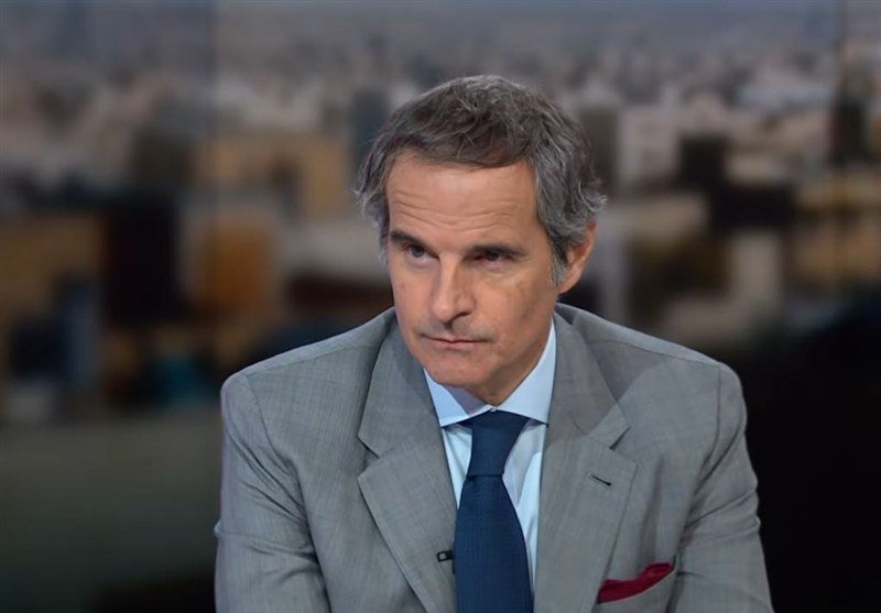 A gray-haired man in a gray suit, blue shirt, and blue tie sits before a blurred cityscape backdrop, looking off-camera with a serious expression.