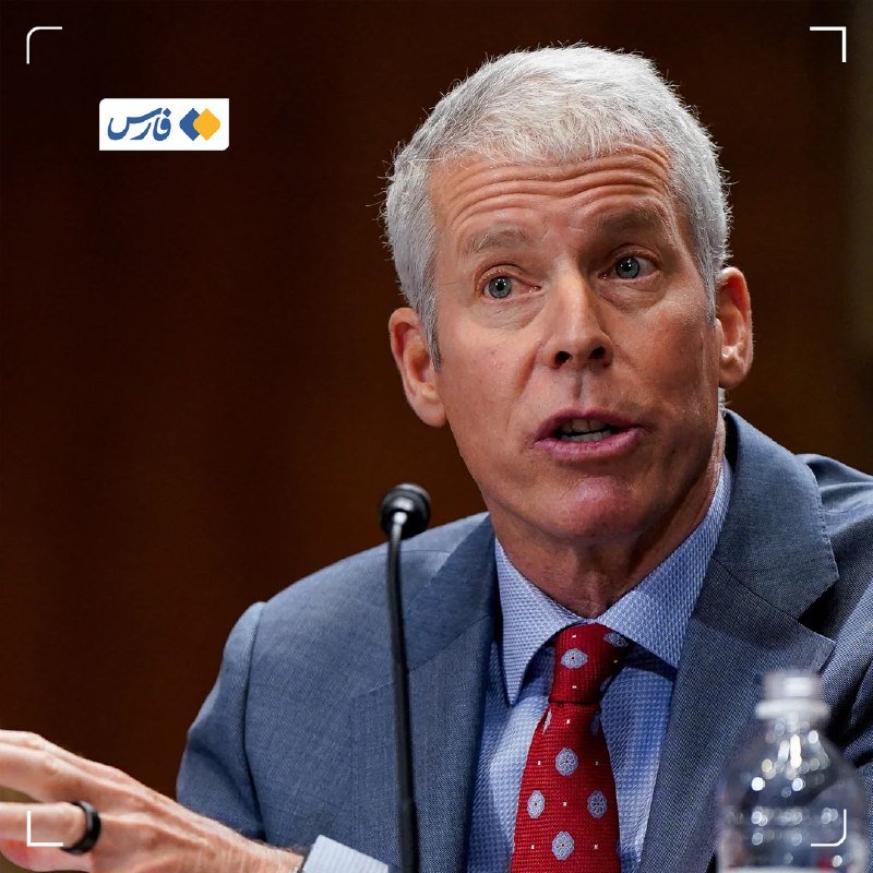 A gray-haired man in a blue suit and red patterned tie speaks into a microphone during what appears to be a formal hearing or testimony.