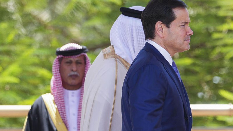 A man in a dark suit walks alongside two men wearing traditional Arab headdresses and robes, with green foliage in the background.