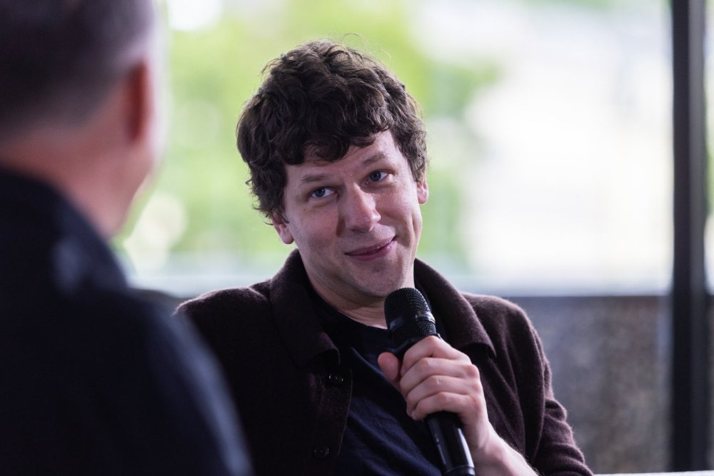 A curly-haired man in a dark jacket smiles while holding a microphone, facing another person visible from behind in the foreground.