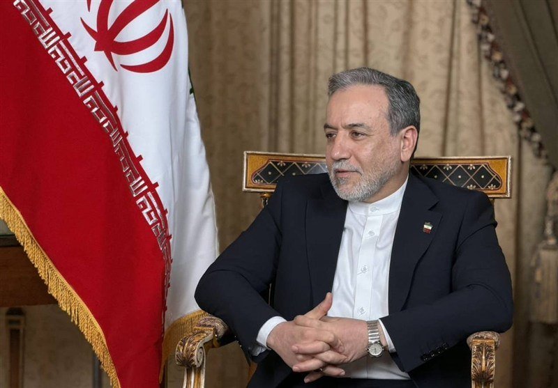 A gray-bearded man in a dark suit sits in an ornate chair beside an Iranian flag.