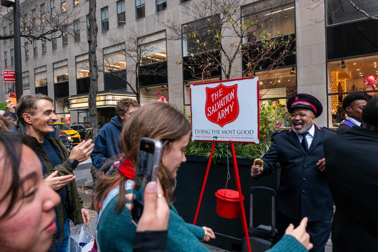 A Salvation Army bell ringer rings a brass bell beside a red donation kettle on a city sidewalk as smiling passersby gather nearby.