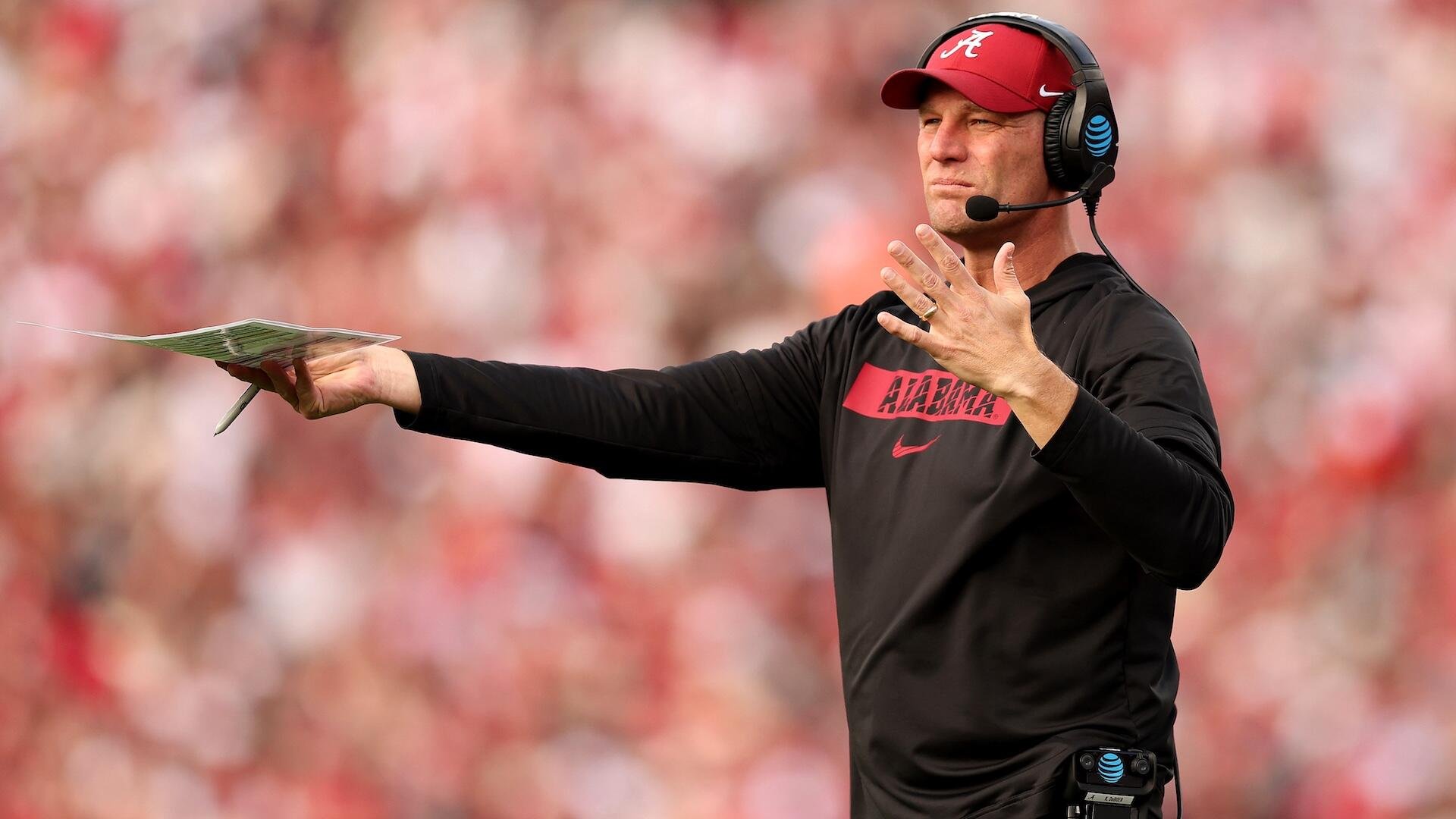 A man wearing a black Alabama shirt, red cap, and headset holds a play sheet and gestures with his other hand on a blurred stadium sideline.