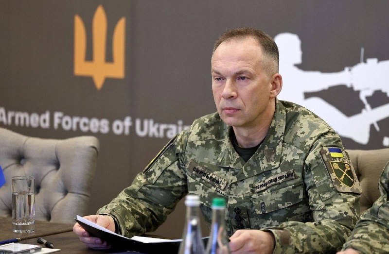 A man in Ukrainian military camouflage uniform sits at a table holding papers, in front of a backdrop displaying the Armed Forces of Ukraine emblem and trident symbol.