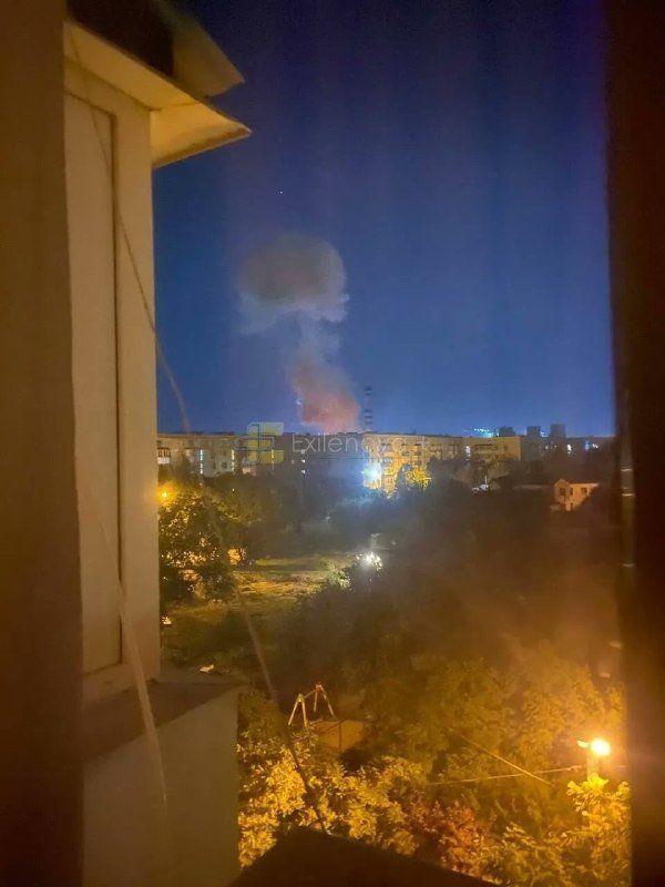 A nighttime cityscape viewed from a window shows a large plume of smoke rising behind illuminated residential buildings.