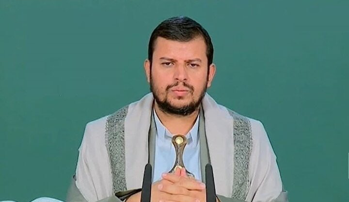 A hand extends holding a stack of Iranian rial banknotes featuring a portrait of a bearded man in a turban, against a blurred gray street background.