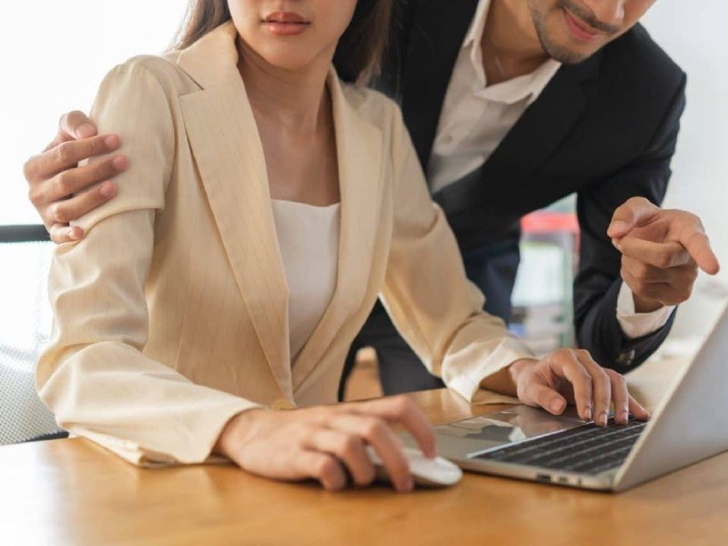 A man in a dark suit stands behind a seated woman in a beige blazer, his hand resting on her shoulder as she uses a laptop and mouse at a desk.