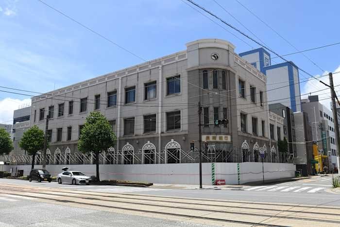 A large, multi-story stone building with arched ground-floor windows stands at a street corner, surrounded by a metal fence, with parked cars and a traffic signal nearby.
