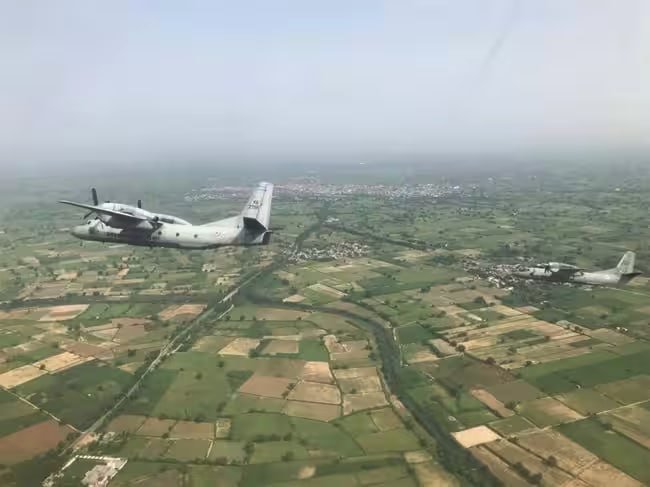 Two military transport aircraft fly in formation over a patchwork of green and tan agricultural fields divided by roads.