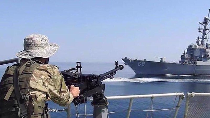 A camouflaged soldier wearing a boonie hat aims a mounted machine gun toward a gray naval warship bearing the number 68, visible on open water in the background.