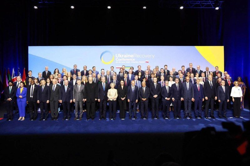 A large group of formally dressed people pose on stage before a blue and yellow backdrop reading "Ukraine Recovery Conference."