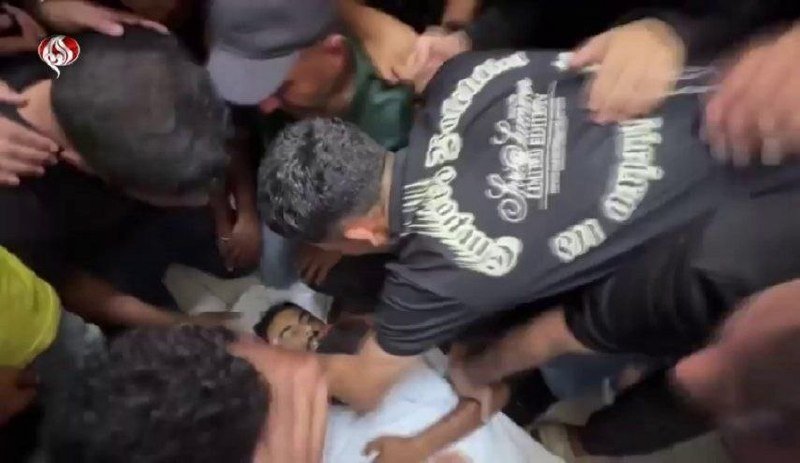 A crowd of people surrounds an injured man lying on the ground, with a man in a black graphic t-shirt leaning over him.