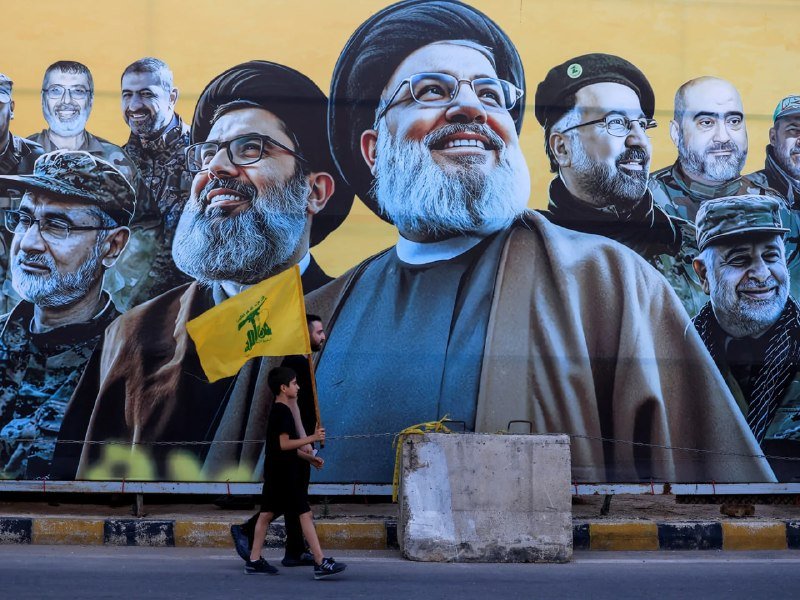 Two boys walk past a large mural depicting multiple smiling, bearded men, with one boy carrying a yellow flag bearing an emblem.