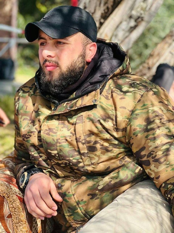 A bearded man wearing a black cap and camouflage jacket sits outdoors near a tree, looking off to the side.