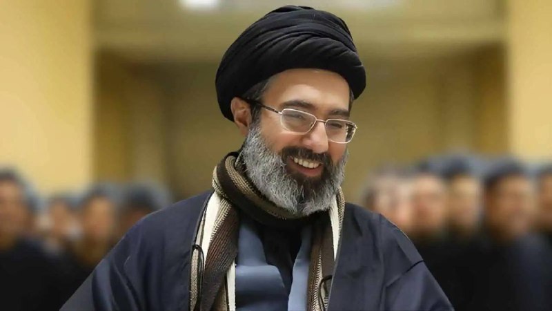 A gray-haired man with a beard, wearing a camouflage military uniform with insignia and an Iranian flag patch, speaks into a microphone.