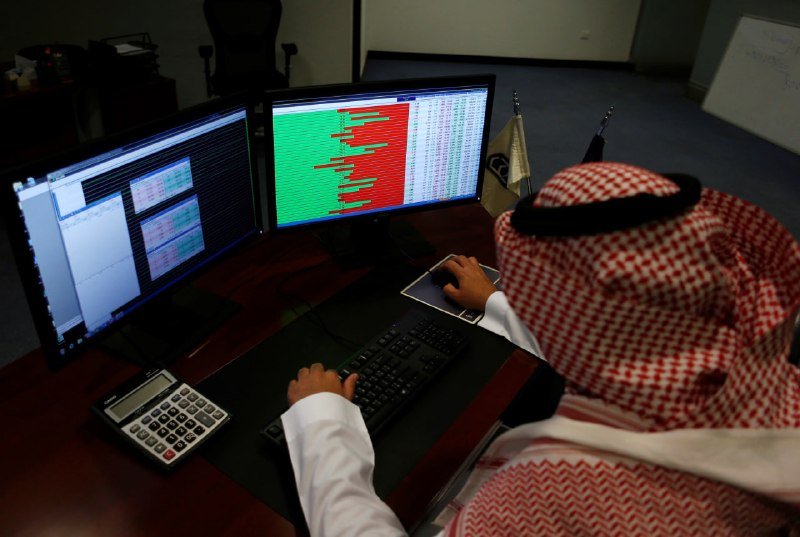 A man wearing a red-and-white checkered keffiyeh and white thobe sits at a desk operating a keyboard and mouse in front of two monitors displaying green-and-red financial charts.
