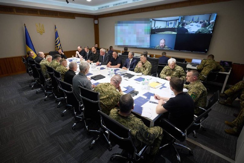 Military personnel and civilians sit around a conference table beneath a Ukrainian flag and coat of arms, with wall screens displaying video feeds of other participants.