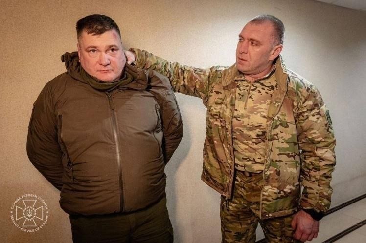 Two men stand against a beige wall; one in a dark jacket, the other in camouflage attire holding the first man's shoulder, with a Ukrainian Security Service emblem in the lower left corner.