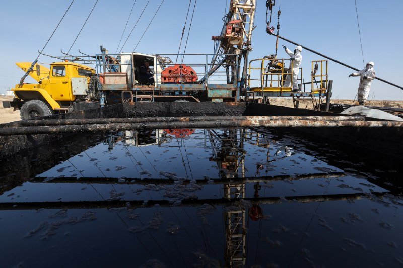Workers in protective gear operate a yellow drilling truck with a tall rig at an industrial site beside a large, dark pool of liquid in an arid landscape.