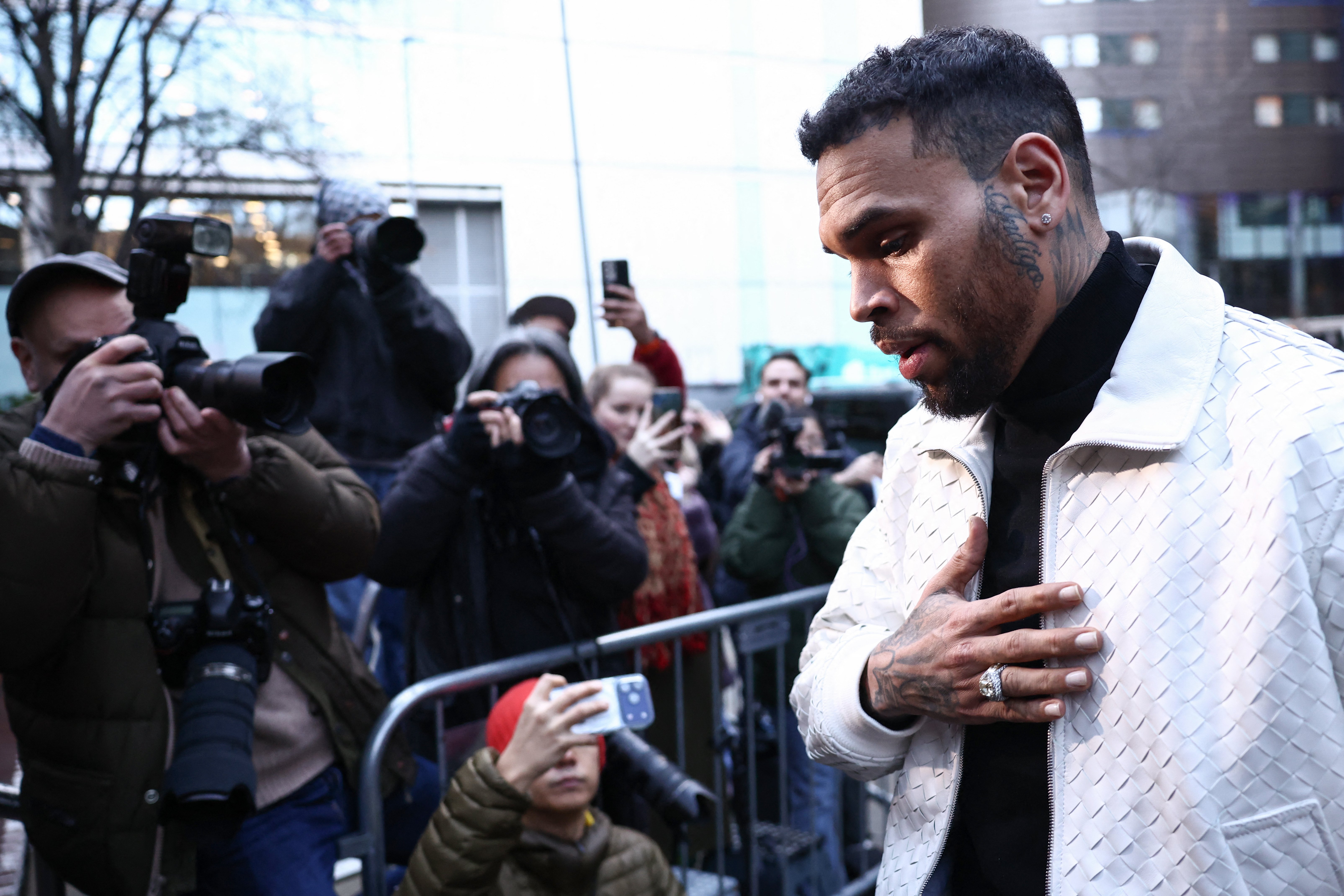 A tattooed man in a white quilted jacket walks past a metal barrier as photographers and onlookers take photos with cameras and phones outdoors.