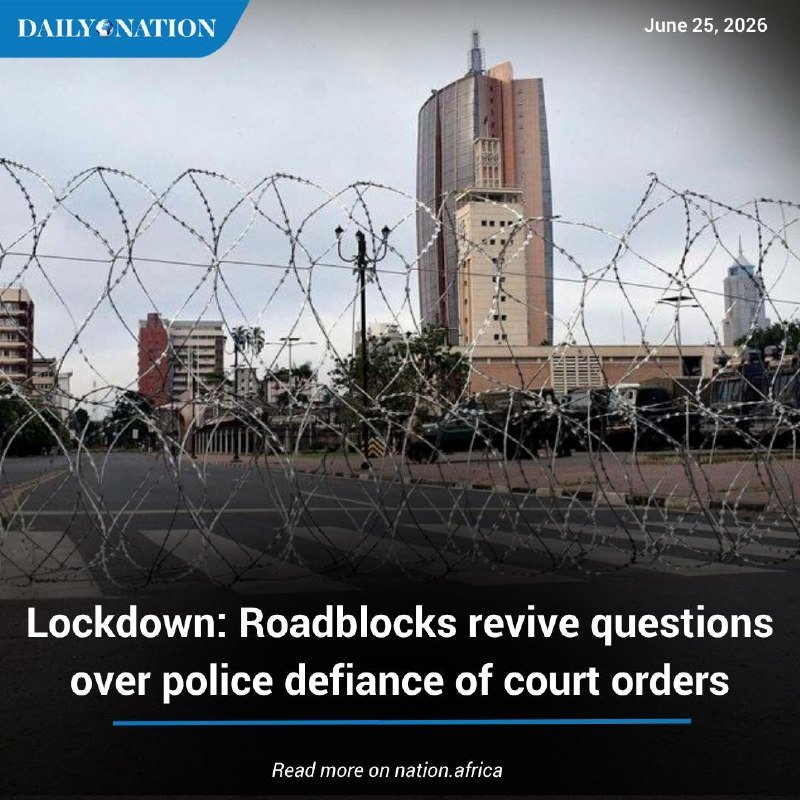 A deserted city street is blocked by coiled razor wire, with a tall tower and buildings visible in the background under an overcast sky.