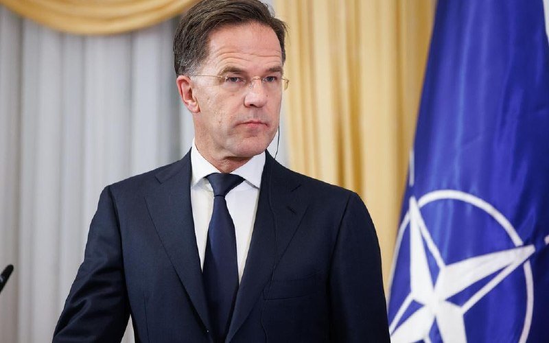 A gray-haired man in a dark suit and tie speaks into multiple microphones labeled "K24," "WDR," "ZDF," and Al Jazeera during a press briefing.