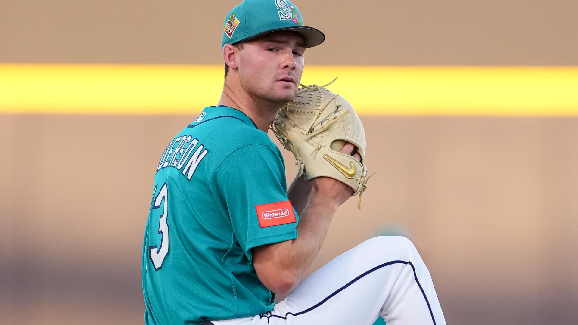 A baseball pitcher in a teal jersey with the number 3 winds up, holding a tan glove near his face during a game.