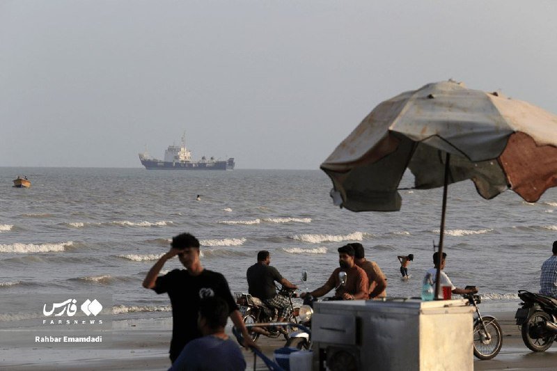 A large cargo ship sails on the ocean horizon while people gather along a hazy beach near a vendor cart beneath a faded umbrella.