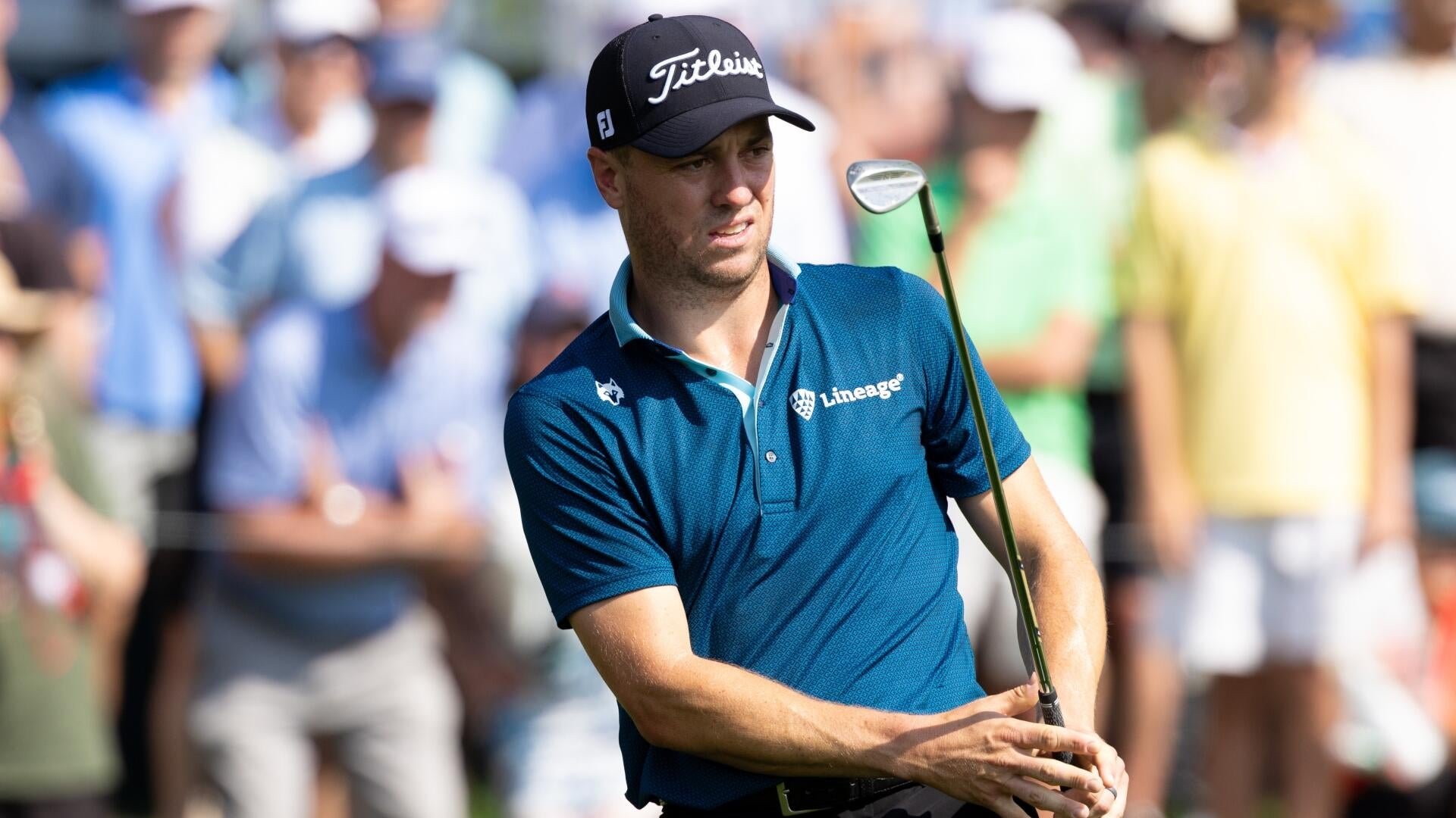 A golfer in a blue Titleist cap and teal Lineage polo watches his shot while holding a wedge, with spectators blurred in the background.