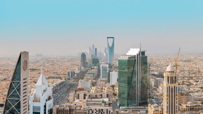Aerial view of a sprawling city skyline featuring multiple modern skyscrapers, including a tall tower with a distinctive arched opening near the top, set against a hazy blue sky.