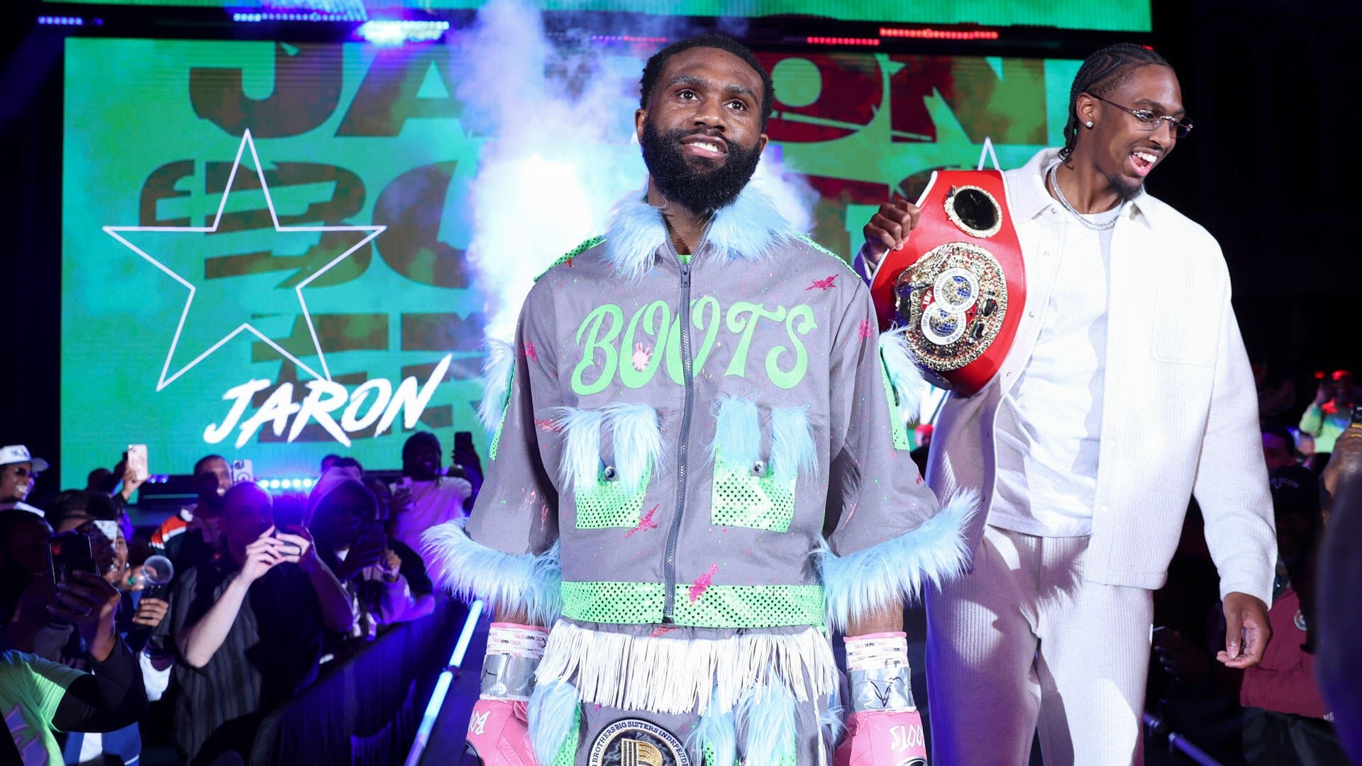 A bearded boxer in a fur-trimmed "BONT'S" jacket walks through a crowd alongside a smiling man in white holding a championship belt, under a screen displaying "JARON" and stars.