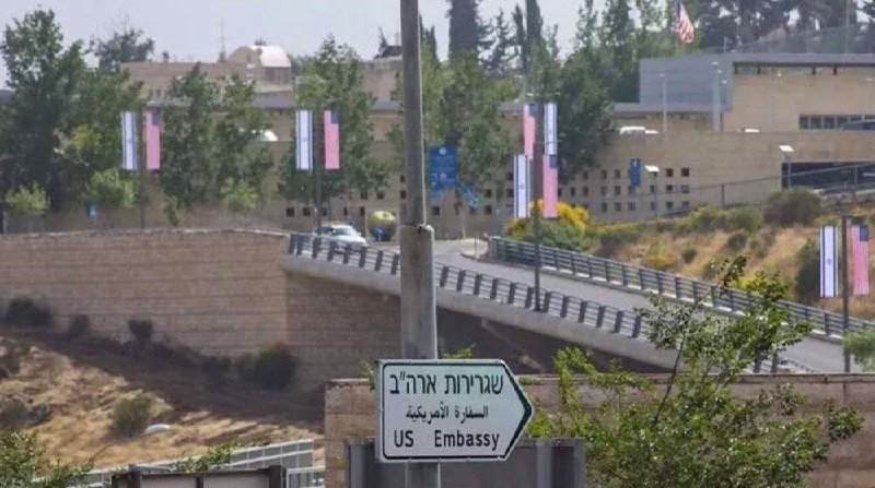 A street sign reading "US Embassy" in English, Hebrew, and Arabic points toward a stone-walled roadway leading to a large building adorned with American flags.
