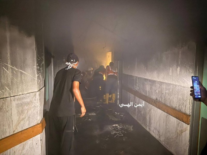 A man in a black shirt walks through a smoke-filled hallway toward a group of people near a fire.