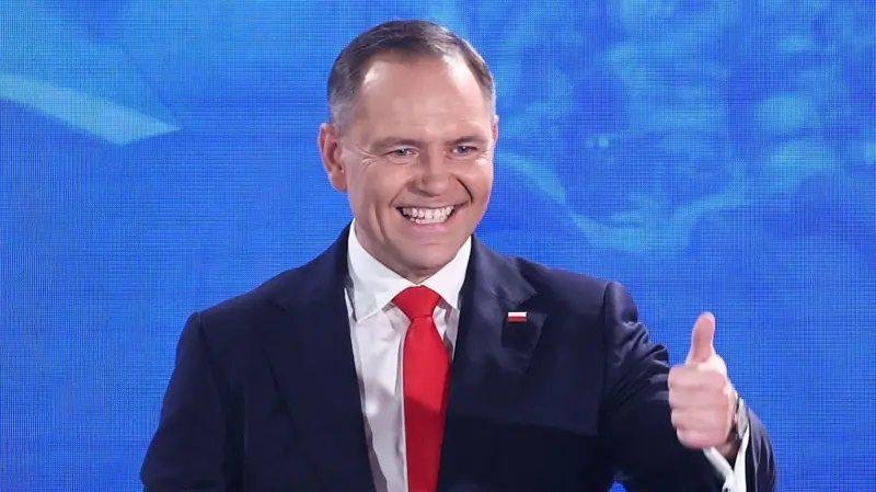 A smiling man in a dark suit, white shirt, and red tie gives a thumbs-up against a blue background.