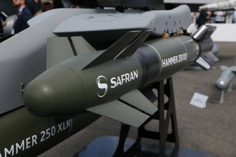 An olive-green Safran "HAMMER 250 XLR" guided munition is displayed on a stand, with blurred attendees visible in the background at what appears to be a defense exhibition.