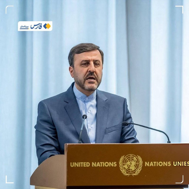 A man with a beard in a dark suit stands speaking at a wooden United Nations podium, with light blue curtains visible behind him.
