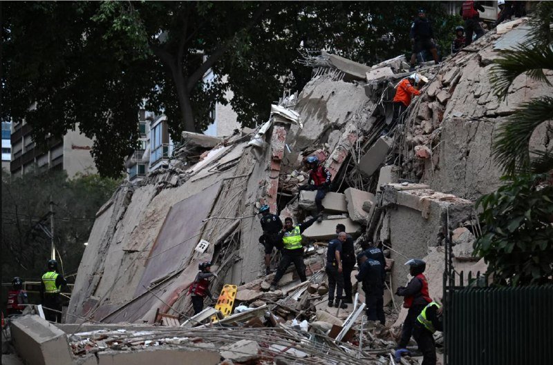 Rescue workers in helmets and high-visibility vests climb over a massive pile of concrete and steel rubble from a collapsed building.