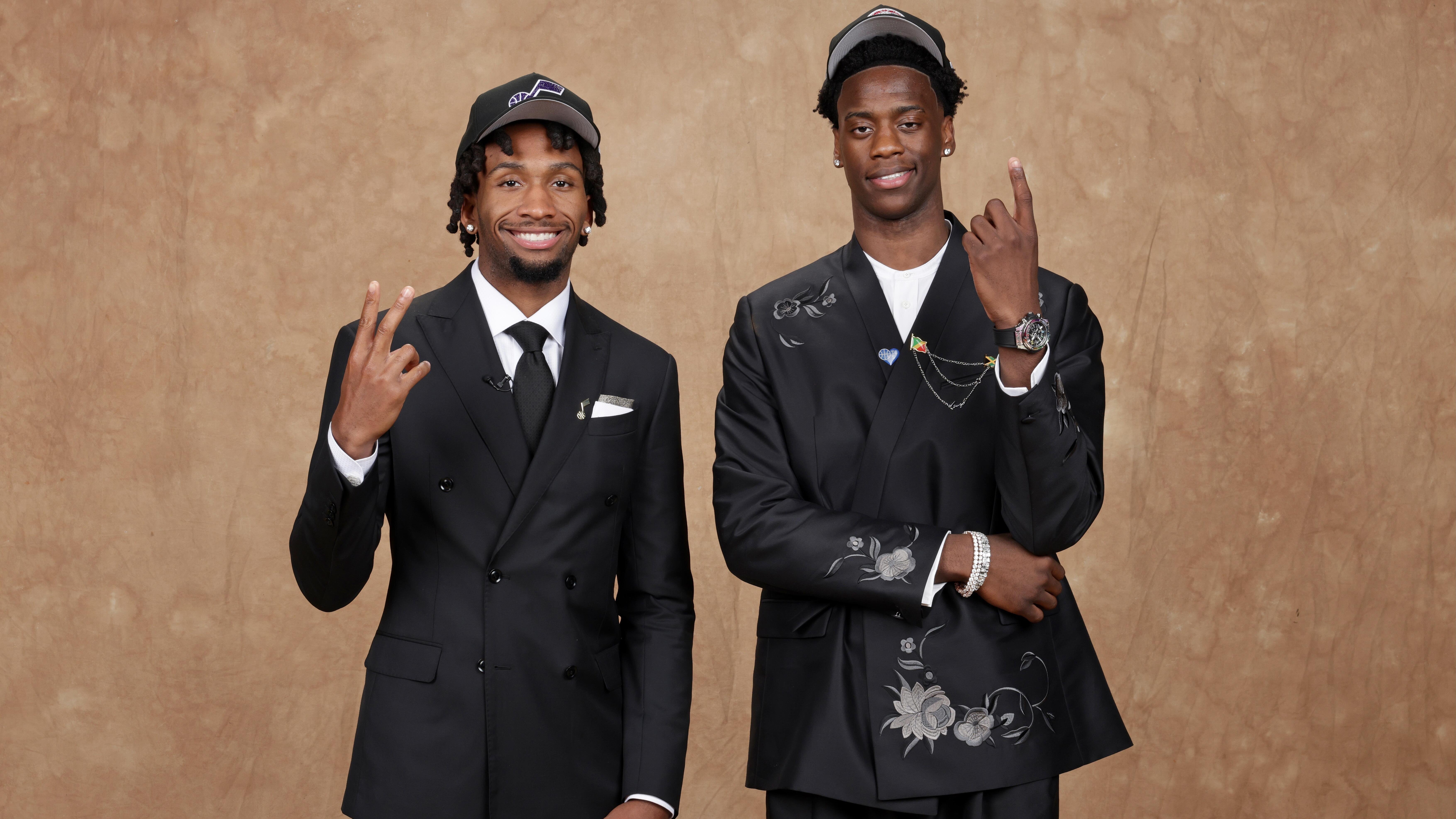 Two young men in black suits and backwards caps pose against a brown backdrop, one giving a peace sign and the other pointing upward.