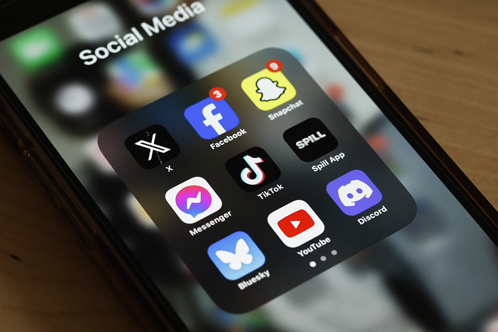 A smartphone resting on a wooden surface displays a "Social Media" app folder containing icons for X, Facebook, Snapchat, Spill App, TikTok, Messenger, YouTube, Discord, and Bluesky.