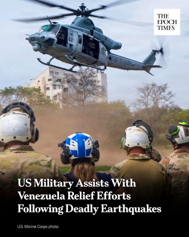 A military helicopter marked "67" hovers low, kicking up dust, as personnel in helmets and headsets watch from the ground, with a damaged building visible in the background.