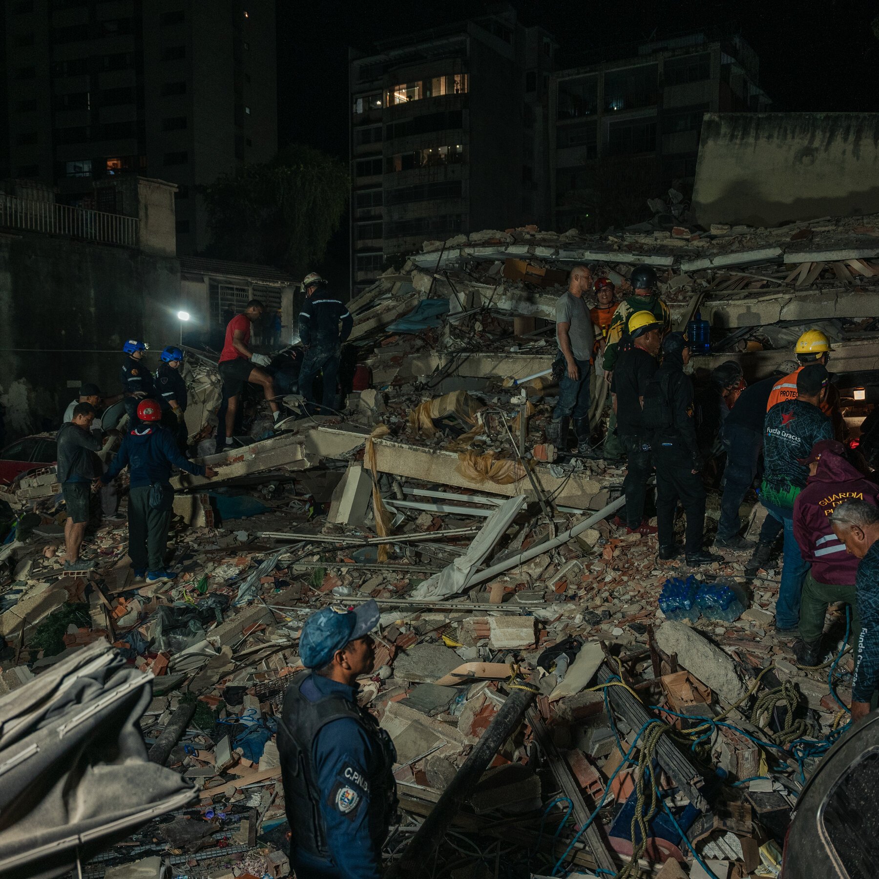 Rescue workers and civilians in helmets search through rubble and debris of a collapsed building at night, illuminated by a single work light.