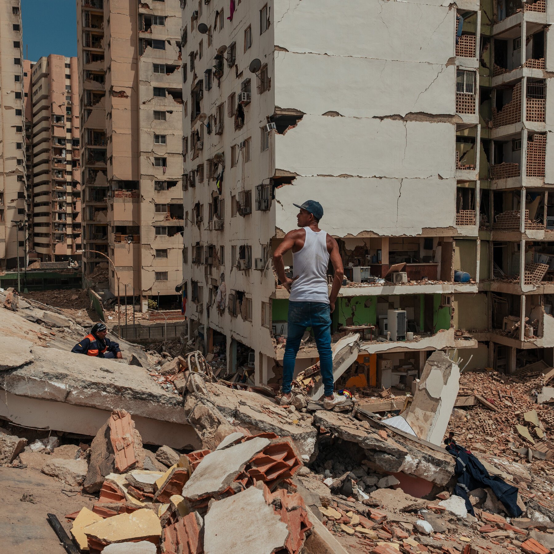A rescue worker in a yellow helmet and blue uniform carries a black and white dog through rubble at night following a building collapse.
