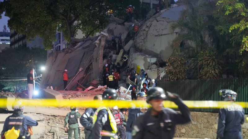 Rescue workers in helmets and uniforms search through the rubble of a collapsed brick building surrounded by palm trees and debris.