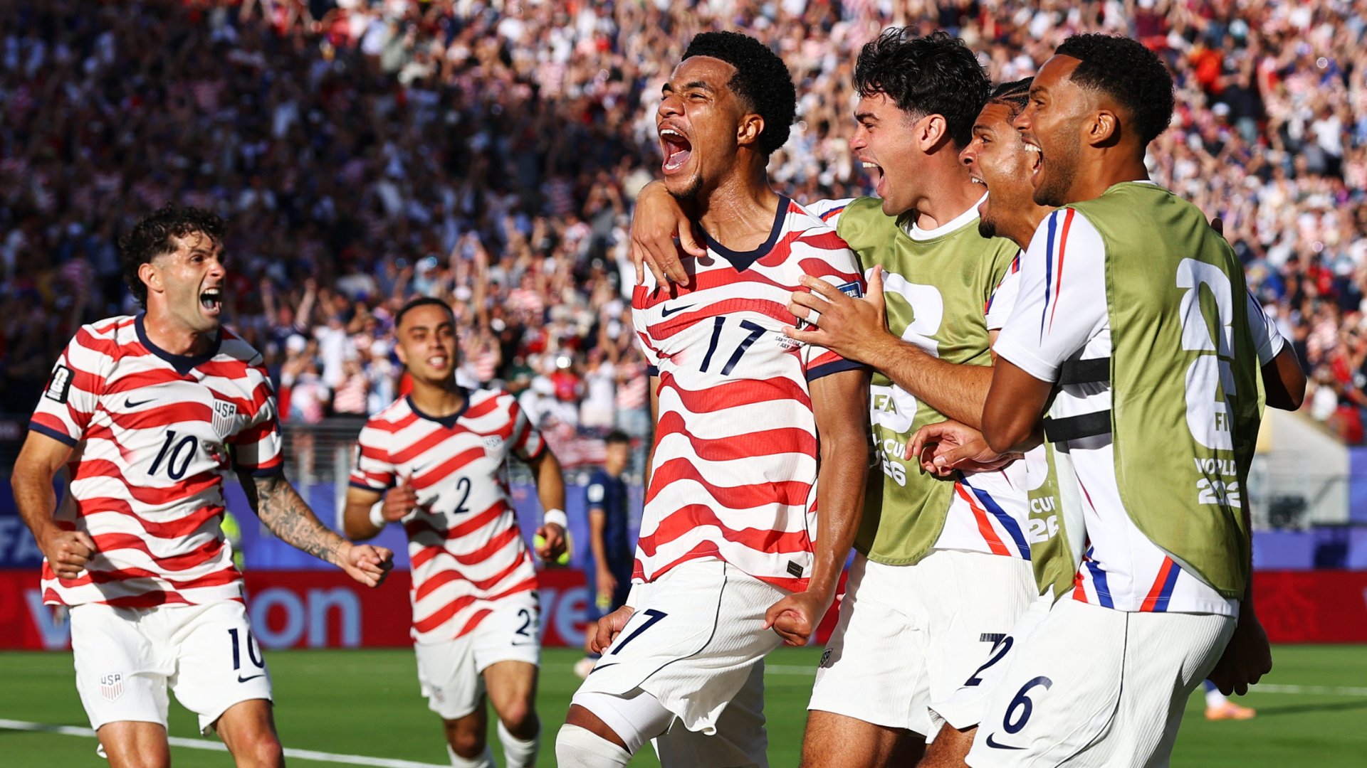Soccer players in red-and-white striped jerseys and gold "FIFA World Cup" bibs celebrate together on a field before a packed stadium crowd.
