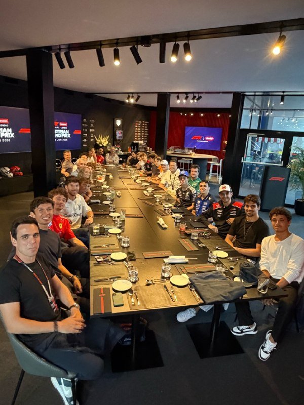 A group of people sit around a long dining table set with plates, glasses, and utensils in a modern venue displaying "Miami Grand Prix" screens.