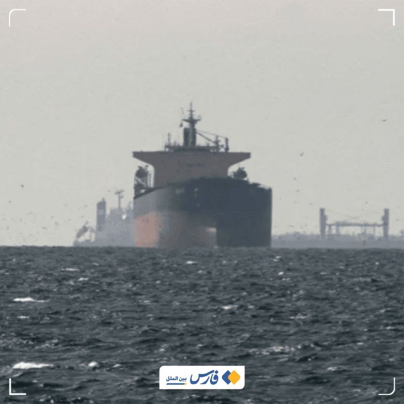 A large cargo ship sails through choppy waters under a hazy sky, with port structures visible in the distant background.