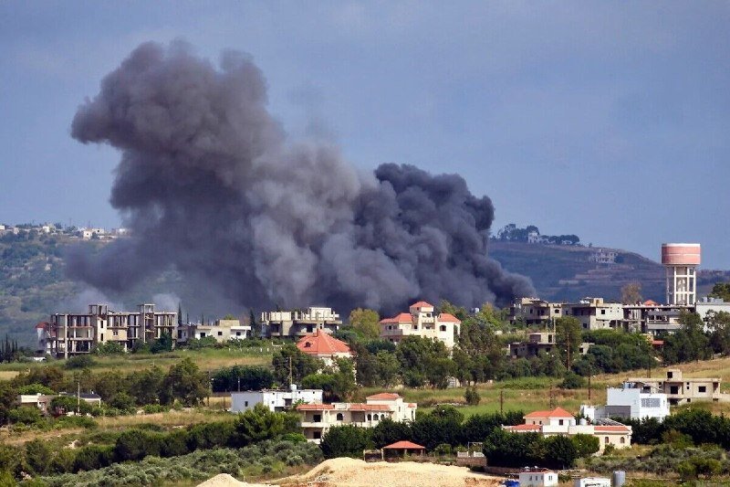 A plume of gray smoke rises above a hilly landscape, viewed past a red-tiled rooftop and distant town under a clear blue sky.