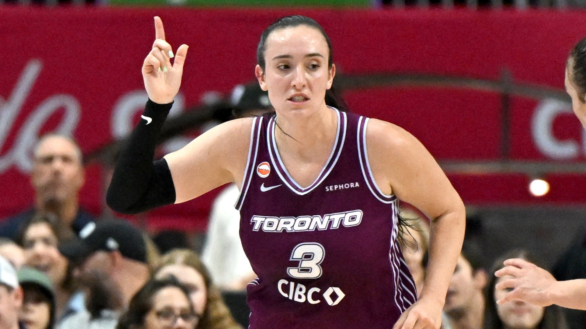A basketball player wearing a maroon "Toronto" jersey with the number 3 raises one finger upward during a game, with spectators visible in the background.