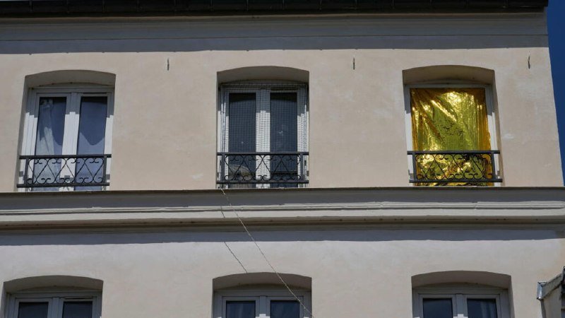 A beige building facade features three upper windows with wrought-iron balconies, one window covered by a shiny gold emergency blanket, while a wire hangs across the lower wall.