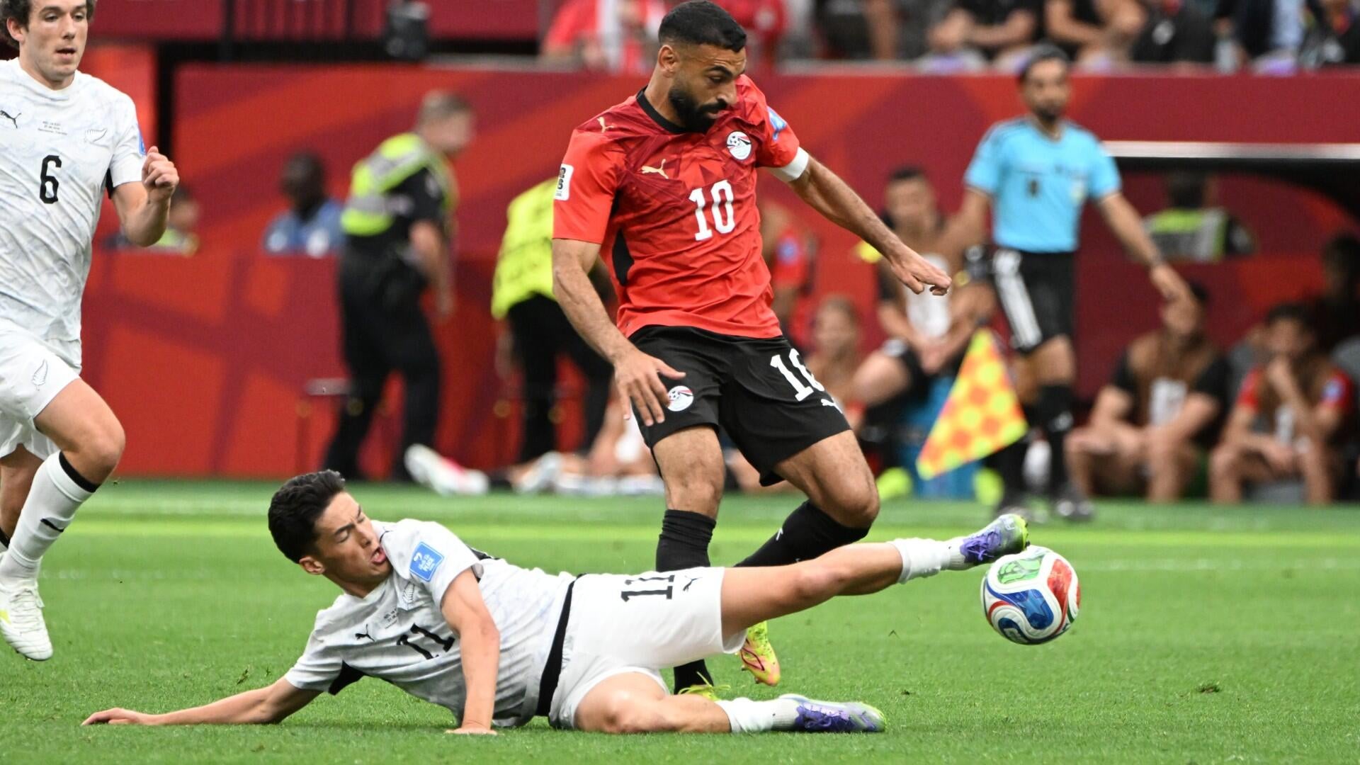 A bearded soccer player wearing a red Egypt FA jersey with a captain's armband stands in a stadium crowd.